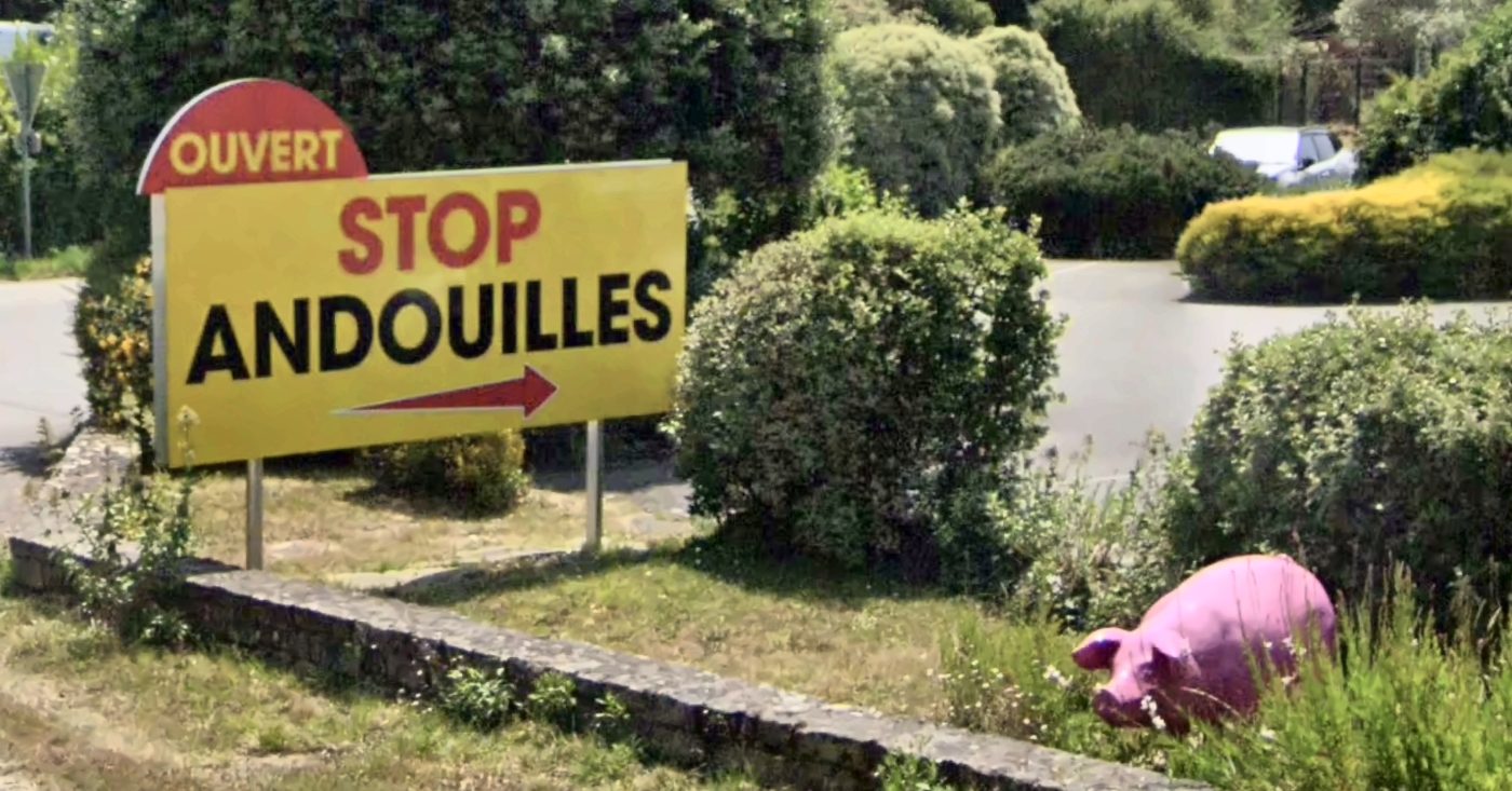 Panneau jaune « Stop Andouilles » au bord d'un rond-point à Plouharnel, avec un cochon en résine dans les massifs